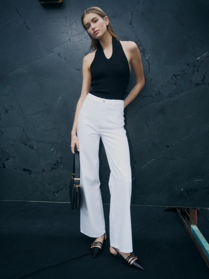Woman in black halter neck knit, white high rise wid leg jean, black michelle flats and black liv mini leather bag